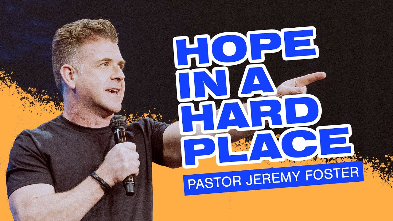 Pastor Jeremy Foster stands with a microphone in his right hand and his left index finger pointing out.