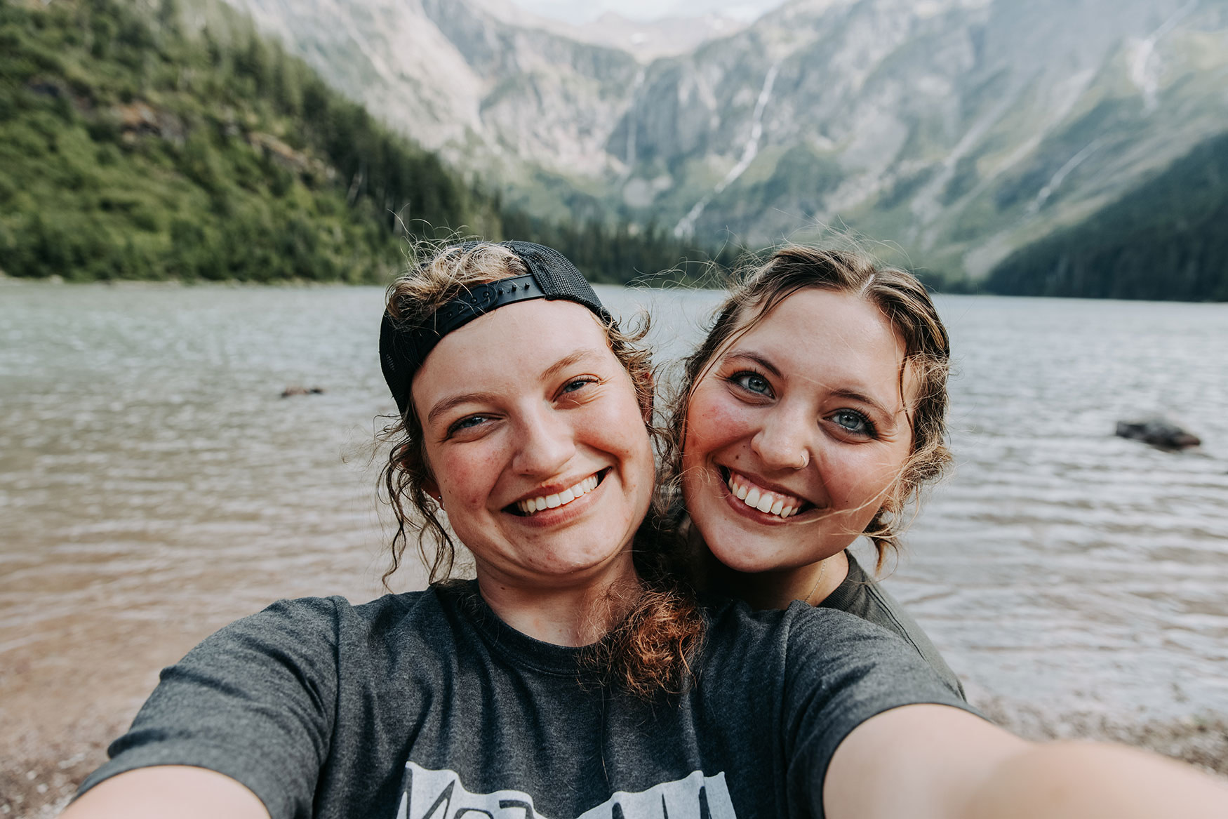 Two girls posting for selfie by a lake and mountains.