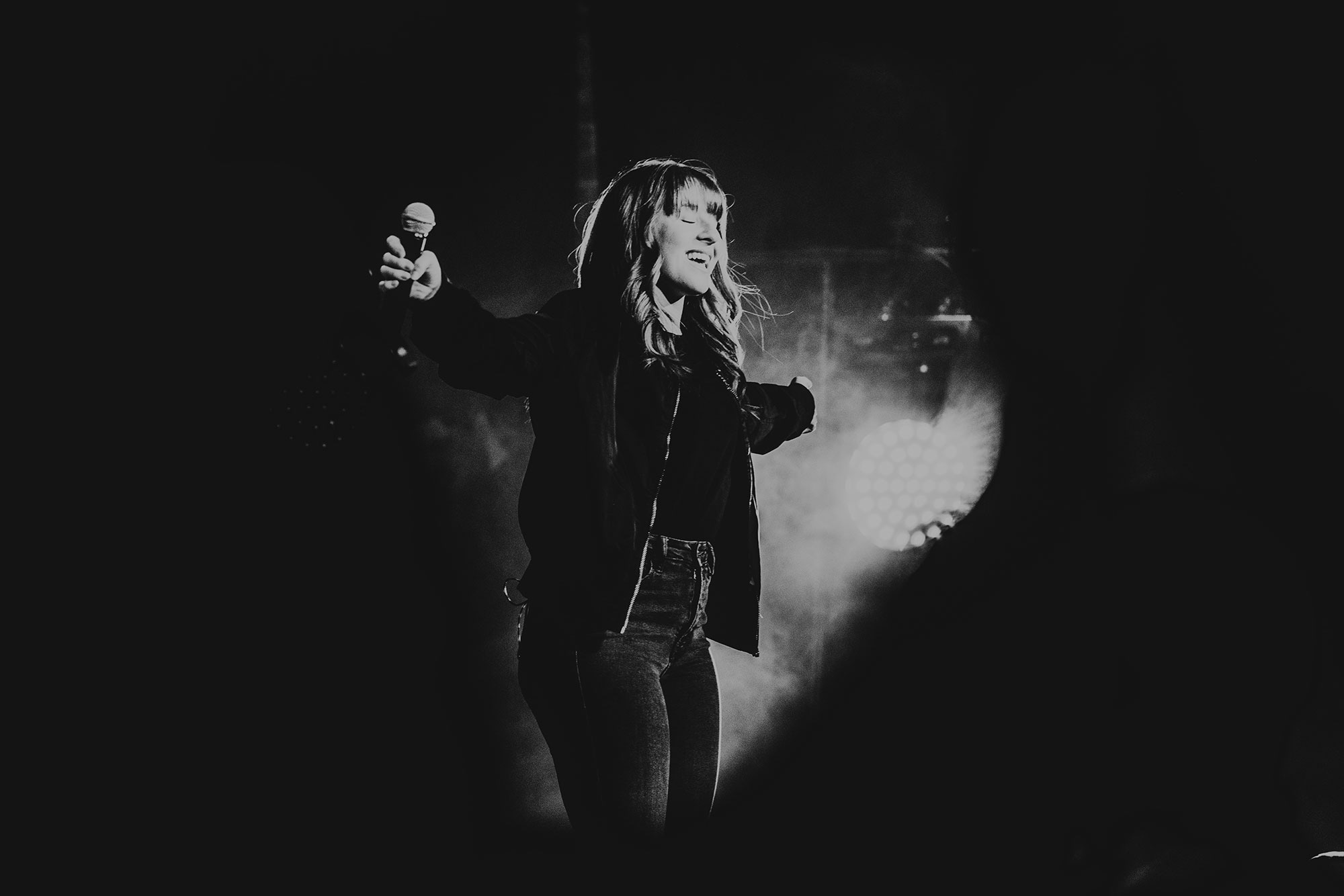 Woman on stage with microphone in outstretch arm in front of a fog machine and lights