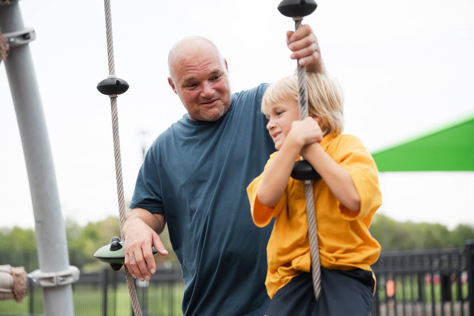 Man helps boy on playground rope swing.