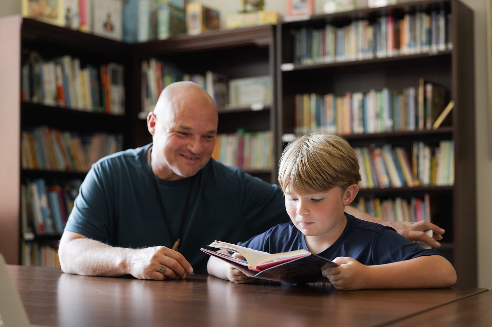 Man helping boy read book in library.