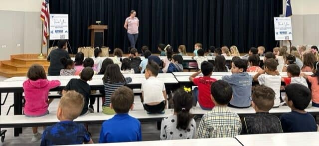 Teacher addressing children in a school auditorium.