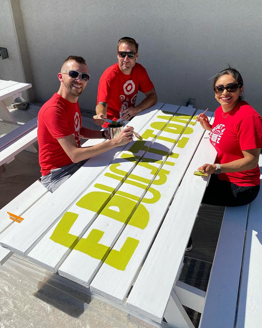Three people painting yellow letters on picnic table.